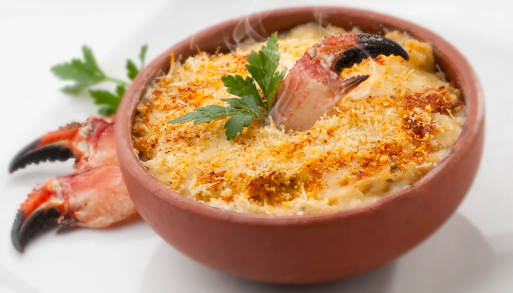 A steaming crab gratin in a clay pot, garnished with parsley and breadcrumbs, with crab claws on the side.