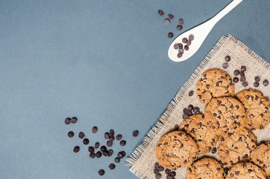 A batch of chocolate chip cookies on burlap, accompanied by scattered chocolate chips and a white spoon on a blue surface.