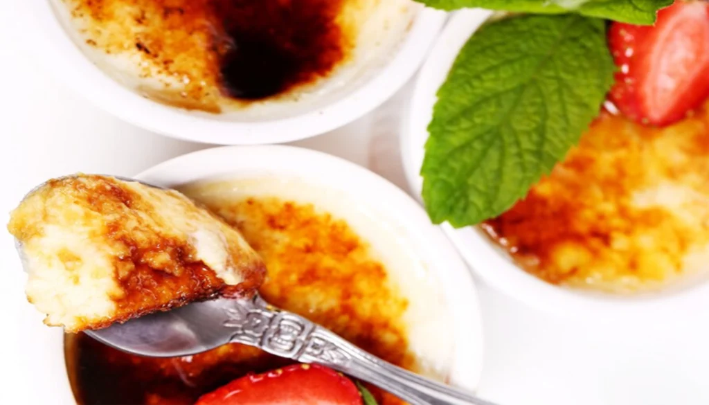 Three ramekins of crème brûlée with caramelized tops, garnished with mint leaves and strawberries.