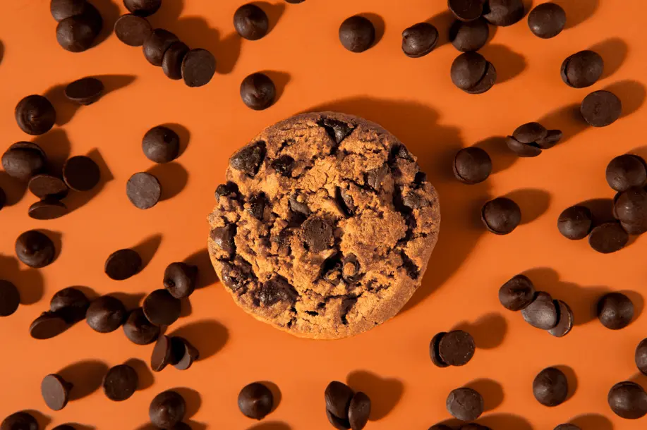 A single chocolate chip cookie surrounded by scattered chocolate chips on an orange background