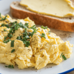 A plate of creamy, fluffy scrambled eggs topped with fresh chopped chives, served alongside a slice of buttered toast on a white plate with a blue rim. Perfect for a quick and delicious breakfast.