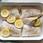 A baking dish with seasoned cod fillets, garnished with fresh lemon slices and a fork resting on one piece.