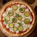 A freshly baked dill pickle pizza topped with Alfredo sauce, garnished with fresh dill on a wooden pizza board.