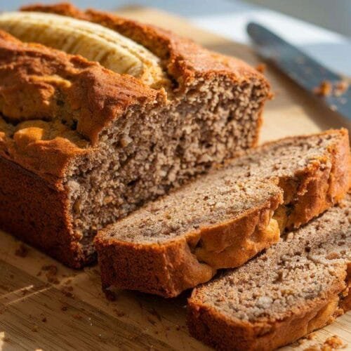 A loaf of golden-brown banana bread sliced to reveal its moist texture, placed on a wooden cutting board with a few slices leaning outward.