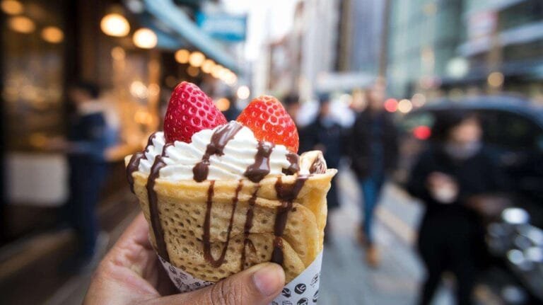 A hand holding a T-Swirl crepe filled with strawberries, whipped cream, and chocolate sauce.