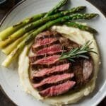 A plated cowboy steak served with mashed potatoes, grilled asparagus, and a garnish of rosemary.